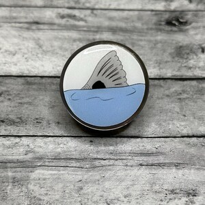 May include: A round, silver-colored pin featuring a gray fish tail emerging from blue water. The background is white, and the pin has a black border. The design is simple and nautical.