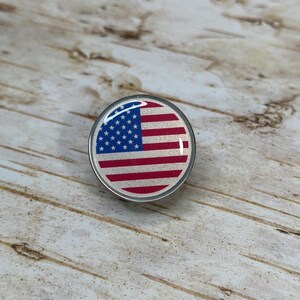 May include: A round, silver-colored pin featuring the American flag design. The flag has red and white stripes and a blue field with white stars. The pin is set against a light wood grain background.