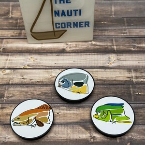 May include: Three round coasters with fish illustrations in shades of green, orange, and gray. The coasters have black edges and white centers. A white block with the text "THE NAUTI CORNER" and a sailboat graphic is in the background.