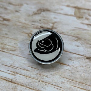 May include: A black and white enamel pin featuring a stylized fish design. The pin is circular and has a silver metal backing.