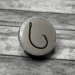 May include: A round, silver-colored object with a black fishing hook design. The hook is a simple, curved line against a textured background. The object is set against a gray wooden surface.