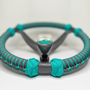 May include: A circular steering wheel spinner knob featuring a gray base and a teal paracord wrap. The handle has a teal and white circular top cap with a graphic design. The paracord is woven in a decorative pattern around the wheel rim.