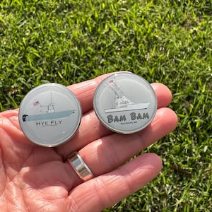 May include: Two round silver metal bottle stoppers with a white background and a boat design. The left stopper has the text "HYE FLY" and the right stopper has the text "BAM BAM" with the text "East Dennis, MA".