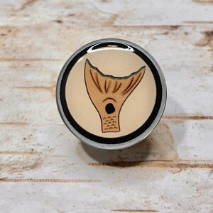 May include: A round, silver-colored bottle stopper with a tan background and a brown mermaid tail design. The tail is outlined in black. The stopper is on a light brown wooden surface.