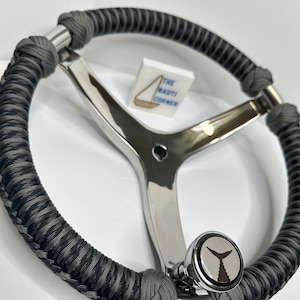May include: A stainless steel boat steering wheel with a gray and black braided grip. The wheel features a three-spoke design and a knob with a fish tail logo. The text "THE NAUTI CORNER" is visible on a small white block.