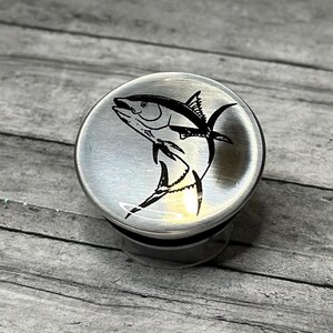 May include: A stainless steel bottle stopper featuring a black and white illustration of a tuna fish. The stopper has a brushed metal finish and a rounded shape, suitable for wine or other bottles.