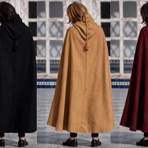 May include: Three hooded capes in black, tan, and burgundy. Each cape has a hood with a tassel and a flowing design. The capes are worn over black pants and shoes. The background features a building with windows.
