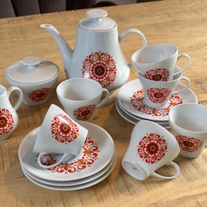 Könnte beinhalten: Weißes Keramik-Teeservice mit einem floralen Design in Rot, Orange und Gelb. Das Set beinhaltet eine Teekanne, eine Zuckerdose, einen Sahnekännchen, Tassen und Untertassen. Das Teeservice steht auf einem Holztisch.