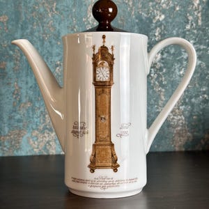 Op de afbeelding: Witte keramische koffiepot met een bruine deksel en handvat. De pot heeft een gedetailleerde illustratie van een staande klok in bruin- en goudtinten. Er staat tekst in het Nederlands op de pot.