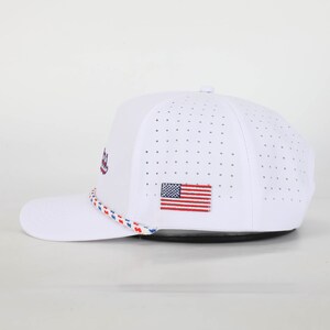 May include: White baseball cap with a perforated design. Features an embroidered American flag patch and a rope detail in red, white, and blue. The cap has a curved brim and a button on top.
