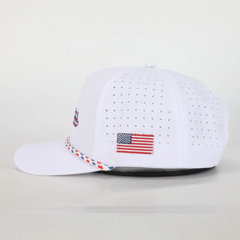 May include: White baseball cap with a perforated design. Features an embroidered American flag patch and a rope detail in red, white, and blue. The cap has a curved brim and a button on top.