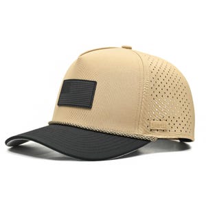 Può includere: Cappellino da baseball beige con visiera nera e toppa nera con bandiera americana. Il cappellino ha un pannello laterale perforato per la traspirazione e un dettaglio a corda sopra la visiera. Il nome del marchio è visibile sul lato.