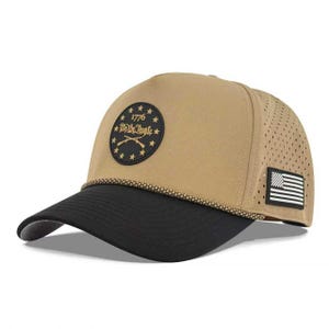 Puede incluir: Una gorra de béisbol color canela con visera negra y un cordón trenzado en la base de la corona. El frente presenta un parche circular negro con estrellas doradas, el texto 1776 y rifles cruzados. Un pequeño parche de bandera estadounidense está al lado.