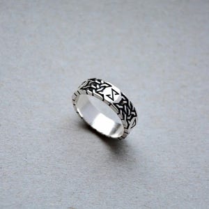 Può includere: Un anello d'argento con un motivo geometrico. L'anello presenta un motivo ripetuto di forme intrecciate attorno alla fascia e un disegno centrale con un simbolo stilizzato. L'anello è mostrato su uno sfondo grigio.
