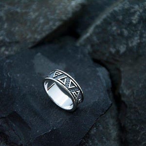Può includere: Un anello d'argento con un motivo geometrico di triangoli e linee. L'anello è posto su uno sfondo scuro e strutturato, che mette in risalto il design dettagliato. La superficie dell'anello riflette la luce, mostrando la sua finitura lucida.