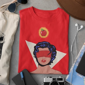 May include: Red t-shirt with a graphic of a woman with a blindfold and an apple with a halo above her head.