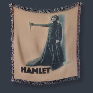 May include: A woven tapestry throw blanket featuring a vintage poster of a theatrical production of Hamlet. The poster depicts a person in a black costume holding a dagger, with the word "HAMLET" printed in black at the bottom.