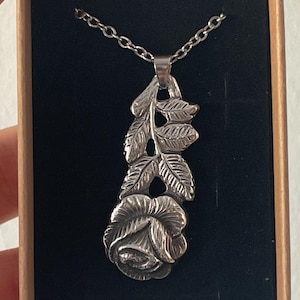 Pu&ograve; includere: Una collana con ciondolo color argento caratterizzata da un design dettagliato di un fiore di rosa con stelo e foglie, sospesa a una catenina sottile. Il gioiello &egrave; presentato all'interno di una scatola di cartone rettangolare con un inserto in velluto nero.
