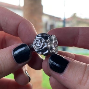 Könnte beinhalten: Ein silberner Ring mit einem detaillierten Rosenmuster. Der Ring wird an einem Finger mit schwarzem Nagellack getragen.