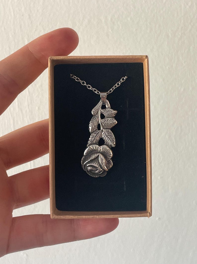 Pu&ograve; includere: Una collana con ciondolo color argento caratterizzata da un design dettagliato di un fiore di rosa con stelo e foglie, sospesa a una catenina sottile. Il gioiello &egrave; presentato all'interno di una scatola di cartone rettangolare con un inserto in velluto nero.
