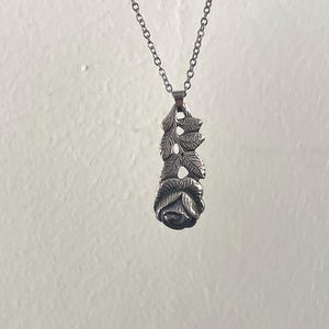 Pu&ograve; includere: Una collana con ciondolo in metallo color argento raffigurante una rosa scolpita alla base con uno stelo verticale e foglie dettagliate che si estendono verso l'alto. Il ciondolo pende da una delicata catena a maglie in metallo color argento.