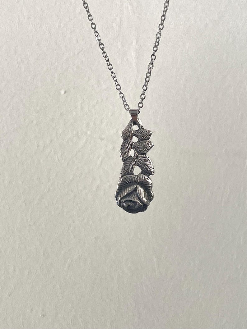 Pu&ograve; includere: Una collana con ciondolo in metallo color argento raffigurante una rosa scolpita alla base con uno stelo verticale e foglie dettagliate che si estendono verso l'alto. Il ciondolo pende da una delicata catena a maglie in metallo color argento.
