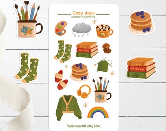 Op de afbeelding: Schattige Cozy Days stickers met illustraties van een regenboog, een kopje warme chocolademelk, een stapel boeken, een paar sokken, een kopje met kwasten, een trui, een bord met pannenkoeken, een wolk met regendruppels en een kopje koffie. De stickers zijn op een witte achtergrond met de tekst "Cozy Days" en "cute stickers by Katerina Arts" bovenaan.
