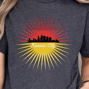 May include: A dark heather grey t-shirt featuring a graphic of the Kansas City skyline within a red and yellow sunburst design. The text "Kansas City" is printed below the skyline.