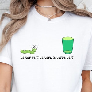 以下が含まれることがあります： 緑の虫と緑のグラスが描かれた白いTシャツ。Tシャツには「Le ver vert va vers le verre vert」という文字が書かれています。