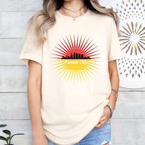 May include: A cream coloured t-shirt with a graphic of a black city skyline with a red and yellow sunburst behind it. The text "Kansas City" is written below the skyline.