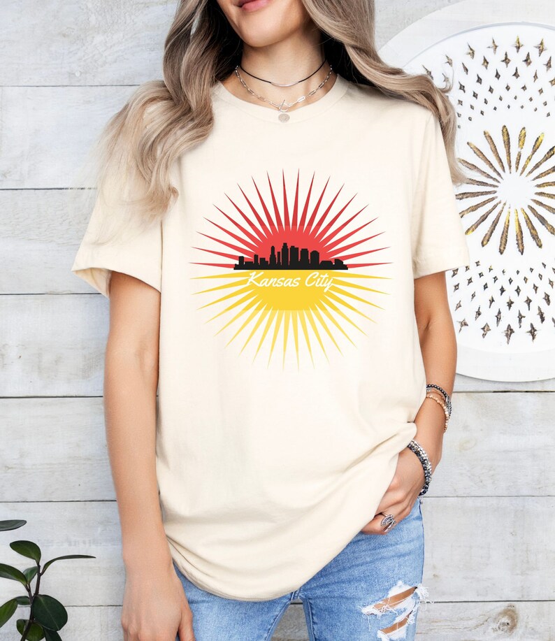 May include: A cream coloured t-shirt with a graphic of a black city skyline with a red and yellow sunburst behind it. The text "Kansas City" is written below the skyline.