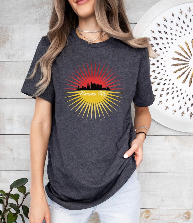 May include: A dark heather grey t-shirt featuring a graphic of the Kansas City skyline within a red and yellow sunburst design. The text "Kansas City" is printed below the skyline.
