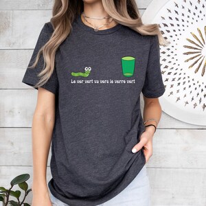 以下が含まれることがあります： 緑色の虫と緑色のグラスが描かれた濃いグレーのTシャツ。Tシャツには「Le ver vert va vers le verre vert.」という文字が書かれています。