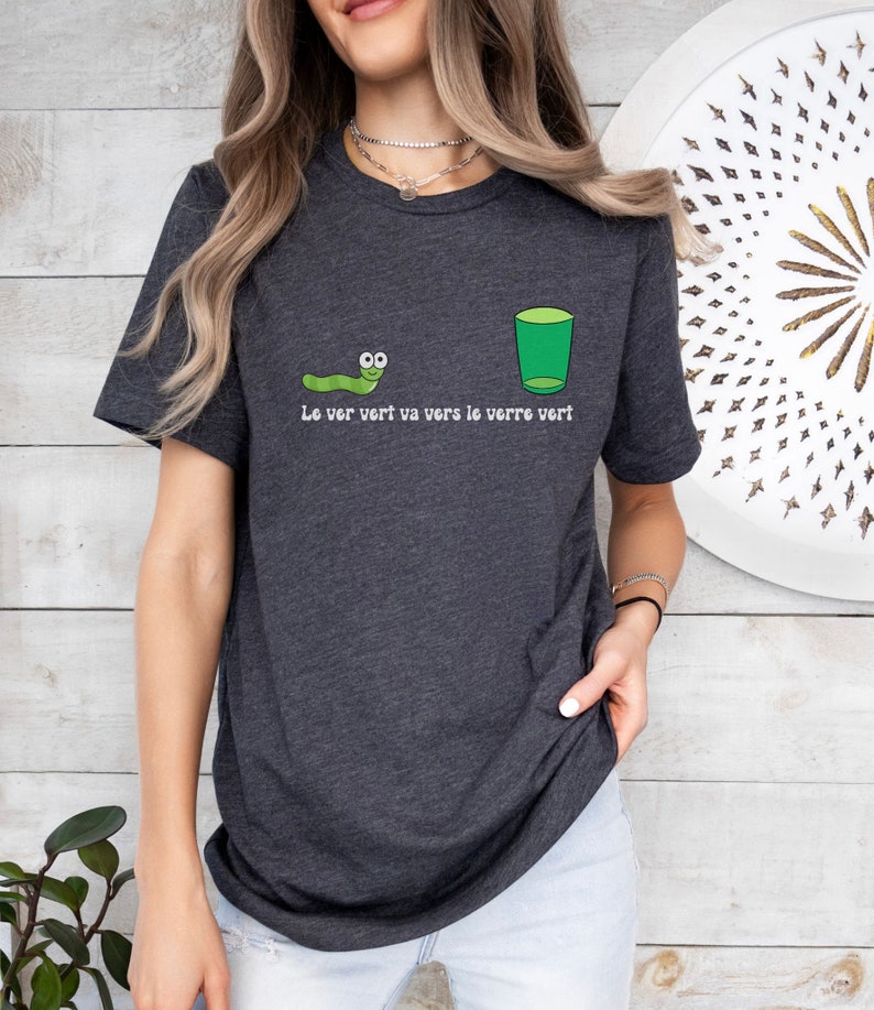 以下が含まれることがあります： 緑色の虫と緑色のグラスが描かれた濃いグレーのTシャツ。Tシャツには「Le ver vert va vers le verre vert.」という文字が書かれています。