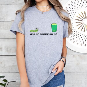 以下が含まれることがあります： 緑の虫と緑のグラスが描かれたグレーのTシャツ。Tシャツには「Le ver vert va vers le verre vert」と書いてあります。