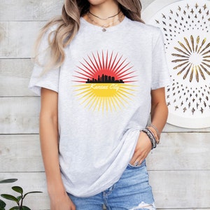 May include: A grey t-shirt featuring a graphic of a red and yellow sunburst with a black silhouette of the Kansas City skyline and the text "Kansas City".