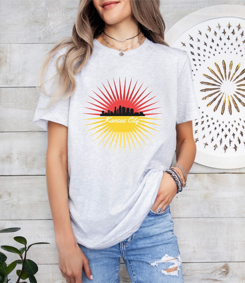 May include: A grey t-shirt featuring a graphic of a red and yellow sunburst with a black silhouette of the Kansas City skyline and the text "Kansas City".