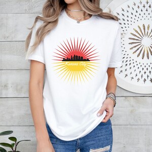 May include: White t-shirt featuring a graphic of the Kansas City skyline in front of a red and yellow sunburst.