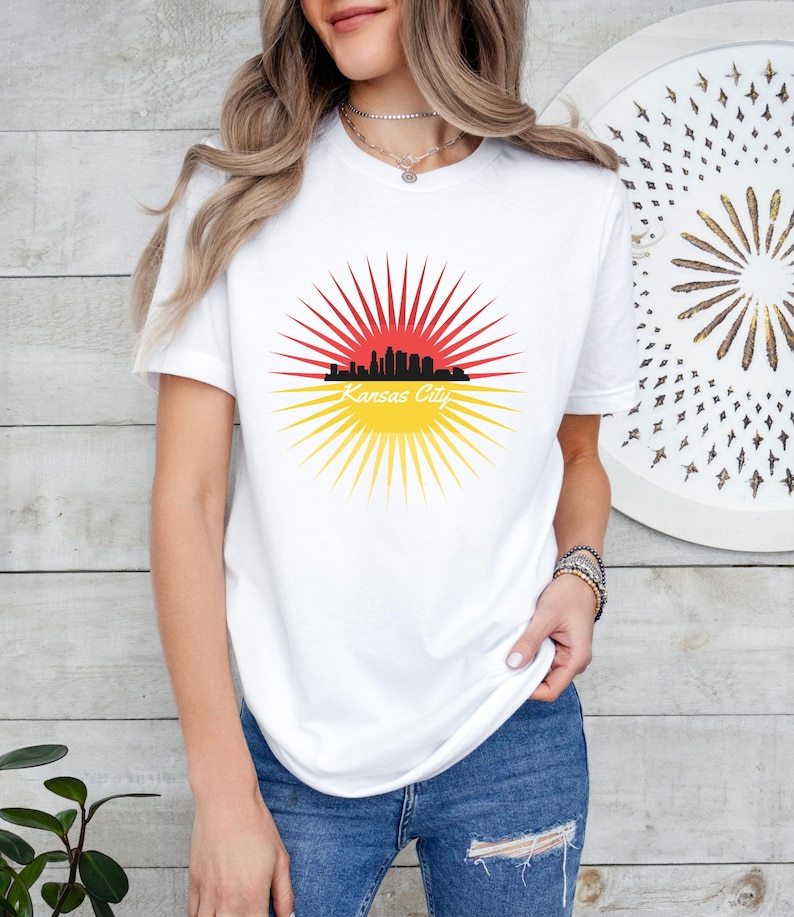 May include: White t-shirt featuring a graphic of the Kansas City skyline in front of a red and yellow sunburst.