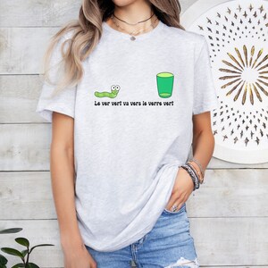 以下が含まれることがあります： 緑色の虫と緑色のグラスが描かれた灰色のTシャツ。Tシャツには「Le ver vert va vers le verre vert」という文字が書かれています。
