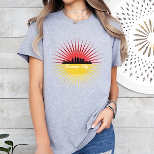 May include: A grey t-shirt featuring a graphic of the Kansas City skyline within a red and yellow sunburst design. The text "Kansas City" is printed beneath the skyline.