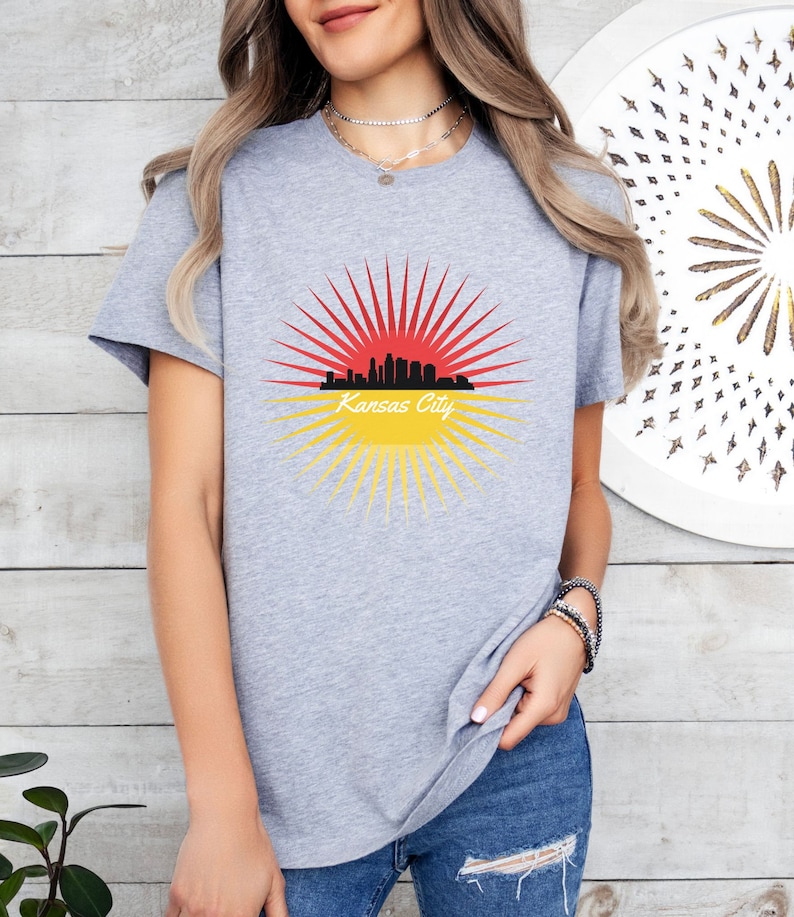 May include: A grey t-shirt featuring a graphic of the Kansas City skyline within a red and yellow sunburst design. The text "Kansas City" is printed beneath the skyline.