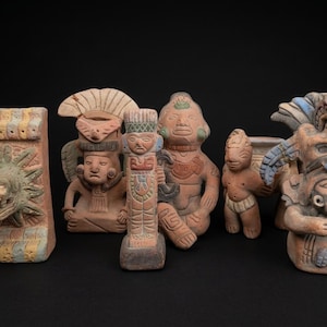 Pode incluir: Uma coleção de estatuetas de terracota retratando figuras maias ou astecas antigas. As estatuetas estão em várias poses e tamanhos, com entalhes detalhados e detalhes pintados em tons de marrom, azul e amarelo. O fundo é preto.