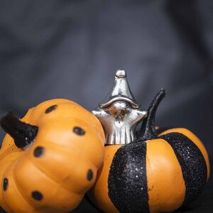 May include: Two decorative pumpkins, one with black polka dots and one with black stripes, are decorated with a silver gnome figurine.