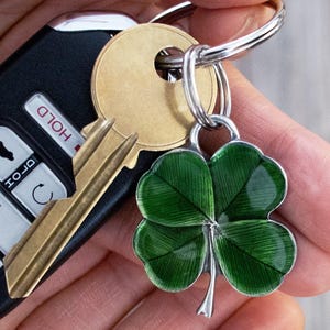 May include: A silver-toned metal keychain charm shaped like a four-leaf clover with deep green enamel inlay featuring fine linear texture. The clover charm is attached to a circular silver split ring along with a brass-colored key and a black car key fob.