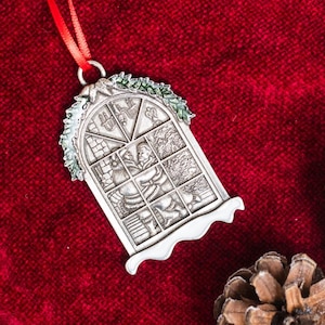 May include: A silver Christmas ornament shaped like a window with a snowy roof and green holly. The window is divided into panes and shows a scene of people inside a house.