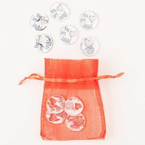 May include: A set of silver-toned metal tokens with the words "You are loved." inscribed on each. The tokens are in a small orange mesh bag.