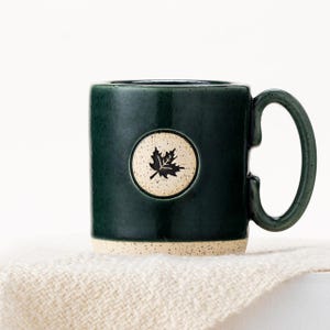 May include: A dark green ceramic mug with a white circle and a black leaf design. The mug has a speckled glaze and a rounded handle.