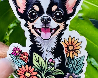 May include: A black and white chihuahua with brown ears is surrounded by colorful flowers. The dog is smiling with its tongue sticking out.