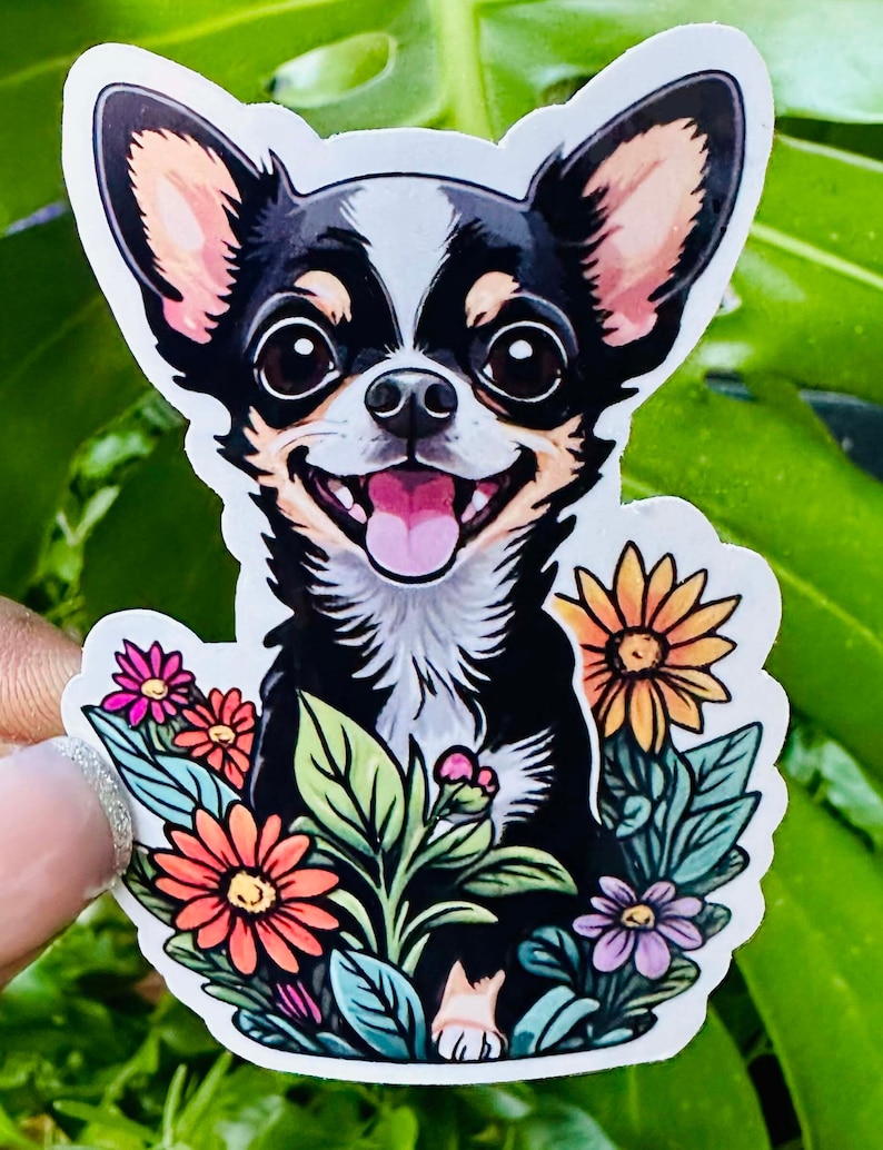 Op de afbeelding: Een zwart-witte chihuahua met bruine oren is omringd door kleurrijke bloemen. De hond glimlacht met zijn tong naar buiten.
